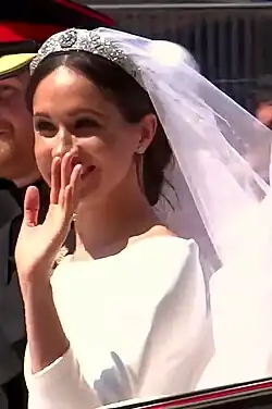 Meghan Markle waves and smiles, wearing a white dress and veil covering her hair. To her left, half out of the photograph, is Prince Harry