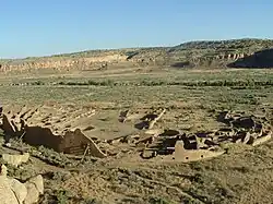 Daytime view looking down on a desert valley: in the near distance, a large semi-circular set of tumbled-down and ruined walls, greyish-yellowish brown in color. The far side of the ruins is a straight line, running left-right, roughly parallel to a line of cliffs in the far distance.