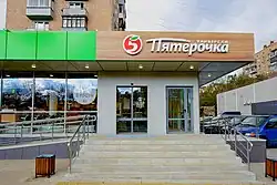 The front of a store, with glass windows and glass doors, and the name of the store in Russian on top, including a logo that appears to be the number five with a leave instead of the upper part of the number.