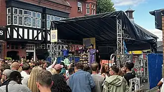 The photo shows a black stage with performers in colourful outfits. There is a black and white pub behind and a large crowd of people below.