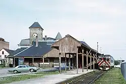 A railcar next to a stone Romanesque railway station with a square clock tower
