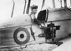 a military member is seated in the observer's cockpit of a Royal Flying Corps plane. The emblem of the Royal Flying Corps is prominently displayed on the side of the aircraft. The military member is seen adjusting a camera mounted to the side of the fuselage, positioned just behind the wing.