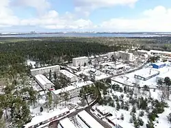 Apartment buildings, detached family homes and several commercial buildings covered in snow in the center, with pines and garages in the foreground, a pine forest stretching on behind the buildings, and a lake in the background