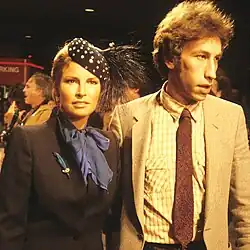 Welch in a blue scarf and high-collared gray jacket, with polka-dot feathered cap, in company of a man