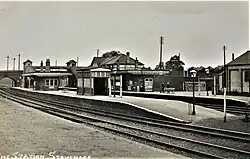 A black and white photograph of a railway station as taken from a short distance away. The station has two island platforms with an additional track passing to the left-hand side. There are buildings on the platforms and a footbridge in the background. Two signs on the platforms say "STEVENAGE", and in the bottom left-hand corner the photograph is captioned "THE STATION. STEVENAGE".