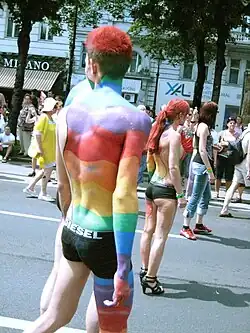 A muscular, young, man walking in a Pride parade in his underwear only and rainbow paint decorating his body.