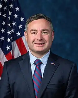 Official House portrait of Vindman smiling in front of the U.S. flag, wearing a black suit with American flag lapel pin, light blue shirt, and striped red and blue tie.