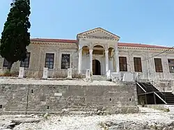 A large, off-white building with a prominent neoclassical porch, held up by columns. A ruined stone wall can be seen in front of it.