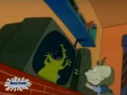 alt=A bald cartoon baby wearing a blue shirt and a diaper stands smiling at a television displaying a large green dinosaur.