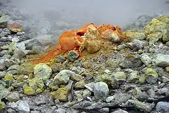 A rocky surface with a mound centrally in the image. The mound has holes in the top. Surrounding rocks have been stained orange and yellow. There is smoke or steam rising from the top.