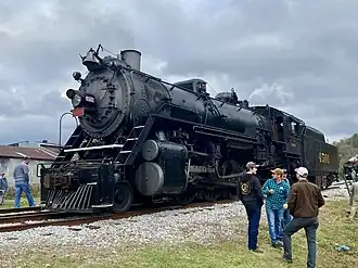 A black steam locomotive with a 2-8-2 wheel arrangement (two leading wheels, eight driving wheels, and two trailing wheels) with tender idling outdoors, surrounded by a crowd of people