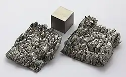 Two samples of rounded dark gray scandium crystals and a metal cube of the same material