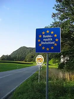A large, square blue road sign in the right-hand verge of a two-lane rural road displays a circle of 12 gold stars with the words "Bundes-republik Deutschland" in white letters inside it. Another smaller, white oval sign a few metres beyond it has the words "Frieslaat Bayern" around a heraldic design.