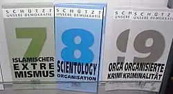 Display with three types of information leaflets, with the numbers and titles "7 Islamischer Extremismus" (coloured in shades of green), "8 Scientology Organisation" (coloured in shades of blue) and "9 Organisierte Kriminalität" (shades of grey). At the top of each leaflet is the inscription "Schützt unsere Demokratie".