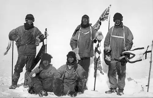 A monochrome image of five men in heavy polar clothing. All look unhappy. The standing men are carrying flagstaffs and a Union Jack flag flies from a half-mast in the background.