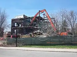 A clawed construction vehicle grabs at an exposed girder in a torn-open building.