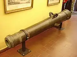 A metal cannon barrel mounted upon two brackets screwed into a brown tiled floor. The cannon barrel is mounted in front of a pale yellow wall supporting two picture frames.