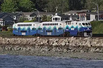 A Sounder trainset, consisting of two double-decker passenger cars and a locomotive, is seen on a track that is slightly elevated over a rocky beach.