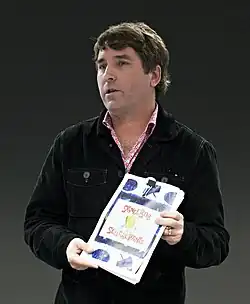 Photograph of Stephen Hillenburg standing holding a book with the title SpongeBob SquarePants looking to his right