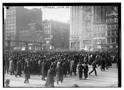 Hundreds of men in black garb and hats holding signs in several languages, flanked by several buildings
