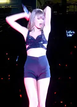 Taylor Swift in a black crop top and shorts, putting her arms on her back, with a microphone on her right arm.