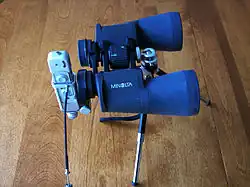 Small, spy sized camera attached to one eyepiece of a set of prismatic binoculars mounted to a tripod on a table top