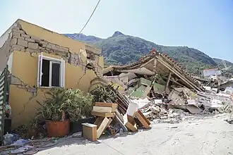 Photo of a small collapsed building with a hill in the background and more collapsed buildings to the right