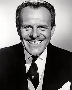 Terry-Thomas, in suit and tie, grins at the camera