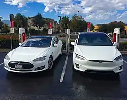 Two white electric vehicles (Teslas) at a supercharger; the one on the left is a sedan while the one on the right is an SUV