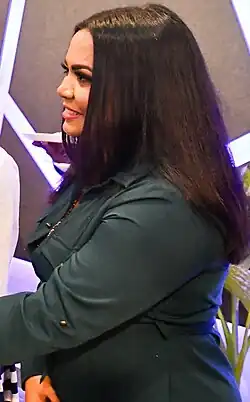 profile image of Thaathi Adam wearing a dark green jacket, face partly obscured by hair, grinning while looking ahead of her