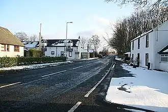 Single carriageway road stretching into distance with houses either side in light snow covered ground on a sunny day with a pale blue sky
