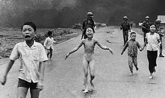 Scared children flee on a road, with soldiers behind them and a smoky sky; in the center is a nude girl, screaming and lifting her arms while running