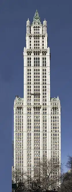 Color photo of the Woolworth Building, a skyscraper, with trees in the foreground and a shorter building to the left