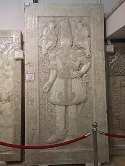 Photograph of a grave stone. Depicted is a monarch in full royal costume with angels on both his top left and top right