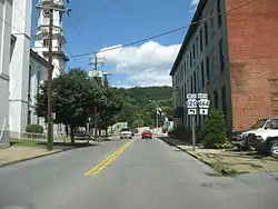 A highway runs toward a bridge between an ornate building with a clock tower and a plain three-story red brick building. A forested hill rises on the far side of the bridge, which is decorated with U.S. flags on poles. A double highway sign along the road indicates that Highway 120 turns to the left (west) before the bridge and that Highway 664 continues across the bridge and heads generally north. Several cars are moving along the highway and over the bridge.
