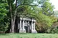 An ornate mausoleum is seen slightly obscured on the left by a tree. It has four columns around its entrance and has TRACY carved above the door. Roof comes to a point, but in layers, giving the impression of a cartoon beehive.
