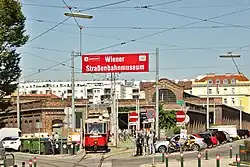 A red historic tram leaving a large brick tram depot, now the Vienna Transport Museum, with a large red sign reading "Wiener Straßenbahnmuseum"