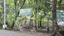 An open entrance to the area of the urban farm designated with a hanging tarpaulin sign, located along a concrete road.