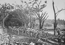 Marines in combat equipment marching along a track which is lined with trees, some of which have been stripped of foliage