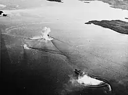 Black and white aerial photo of two ships sailing together near the shoreline. One of the ships is partially obscured by a large splash.