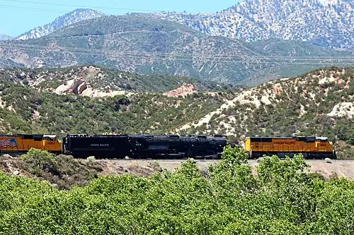 Two yellow diesel locomotives hauling an articulated steam locomotive, one at the front and the other at the back, through the mountains in the background