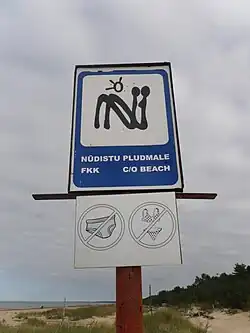 Blue sign with styilised N, reading "Nudistu pludmale fkk C/O beach", and pictures of swimsuits with a strikethrough.