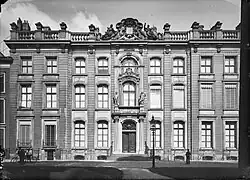 The façade of a neoclassical building; on the left are two men with bicycles and on the right is a man on foot