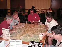 Five men sit at a table, poring over a map. Dice and counters lie atop the map, some counters are stacked. The map is subdivided into small hexagons of identical size.