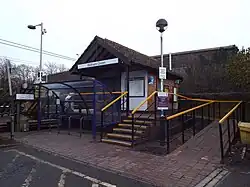A tiny brick structure with stairs and a ramp leading up to it. A canopy covers the path in front of the building, with a sign on the canopy that reads "Welham Green". In the left-hand background overhead wires are visible behind a fence.