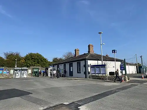 Photo of entrance of Wicklow Train Station taken from outside. The picture is taken during the day. The sky is blue, with white clouds. There are cars parked outside the station entrance. The station office (on the right hand side) is a yellow building (one storey) with green borders. There are trees on the background. The foreground is the road where the photo is taken from