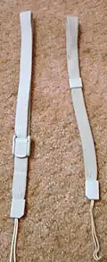A close up photograph of two light-grey wrist straps laid parallel and vertically on beige carpet. The left strap is slightly thicker compared to the right strap. Both straps have a sliding plastic keepers to adjust the size and tension of the strap's loop; the left keeper includes a simple locking mechanism that is absent from the right keeper.