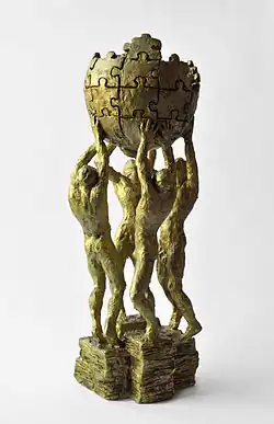 A small model of the sculpture. It depicts four people holding up an incomplete sphere made of jigsaw puzzle pieces.