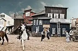 Background: Facades of Wild West themed buildings. A man is riding a horse and people are running in the background.