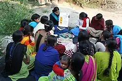 A woman teaches women's literacy class. There is a group of women surrounding a single woman who holds up a board on which there is foreign writing. She is teaching literacy to the group of women.